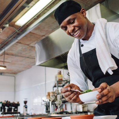 Gourmet chef in uniform cooking in a commercial kitchen. Happy male cook wearing apron standing by kitchen counter preparing food.
